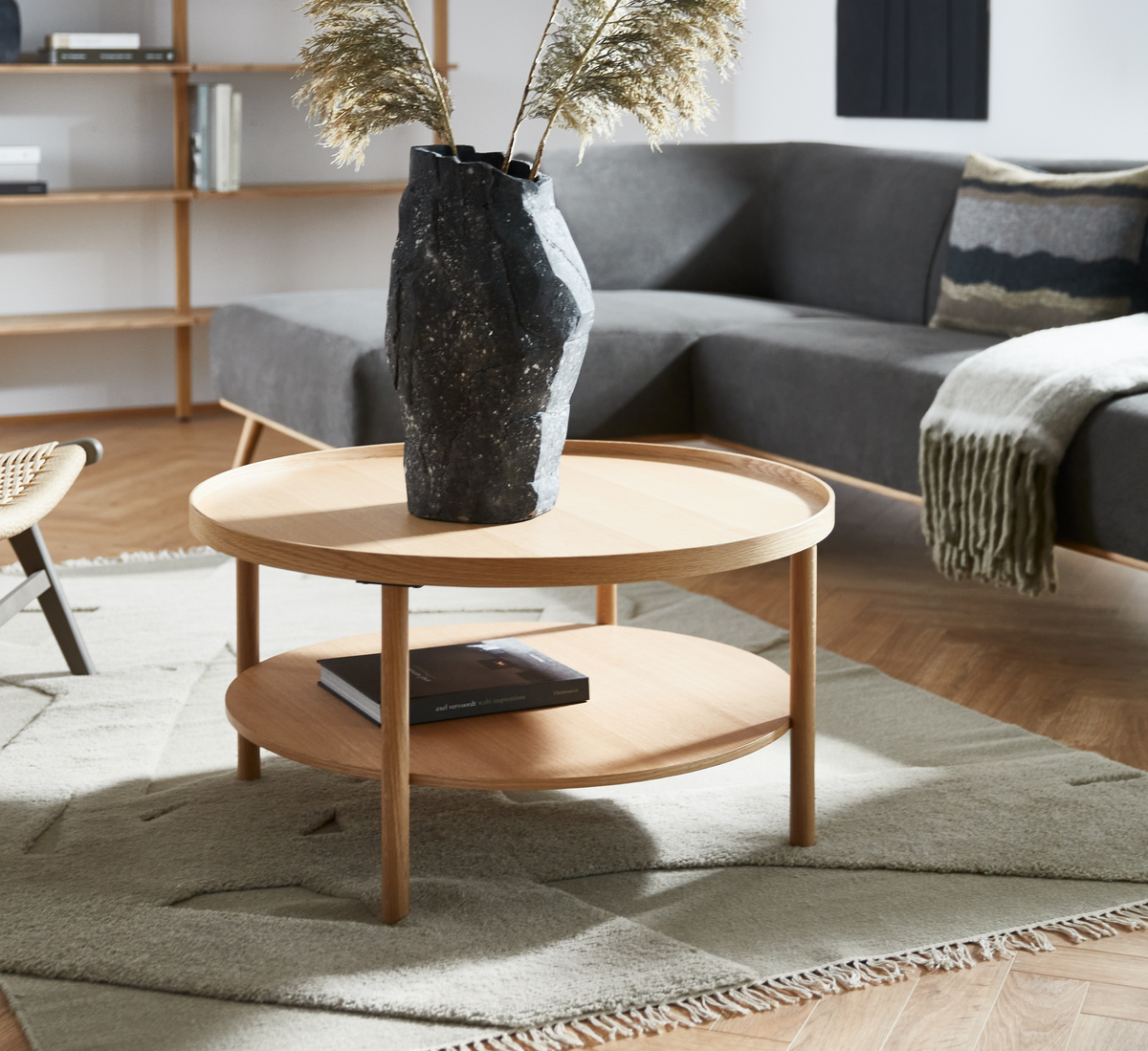 Rundt sofabord i lys eg med hylde, sort vase med tørrede strå og en bog; grå sofa og lyst tæppe i en moderne stue.