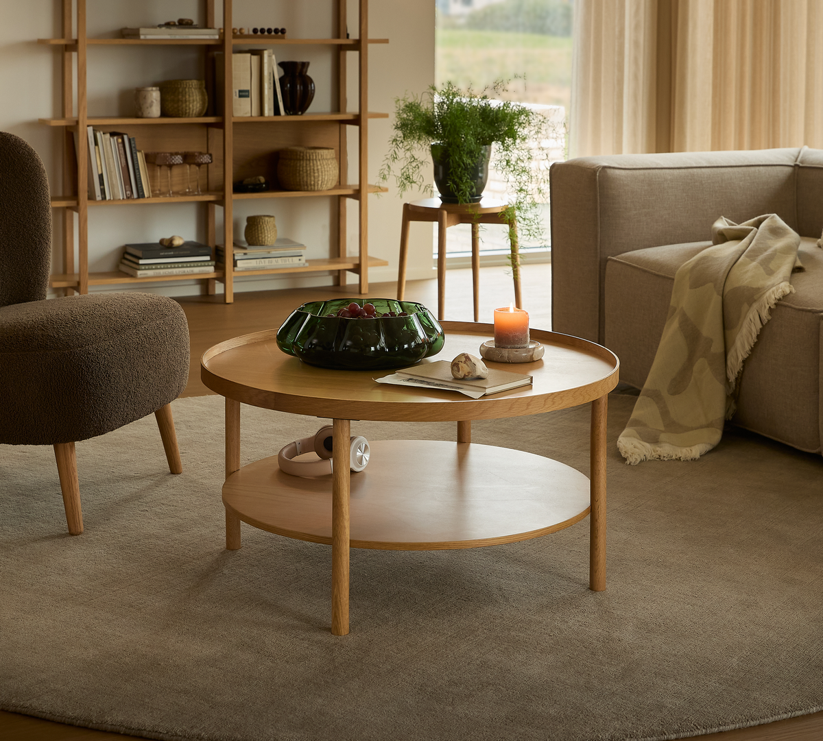 Rundt sofabord i eg med hylde, pyntet med grøn glasskål, tændt lys og bog i en varm, skandinavisk stue.