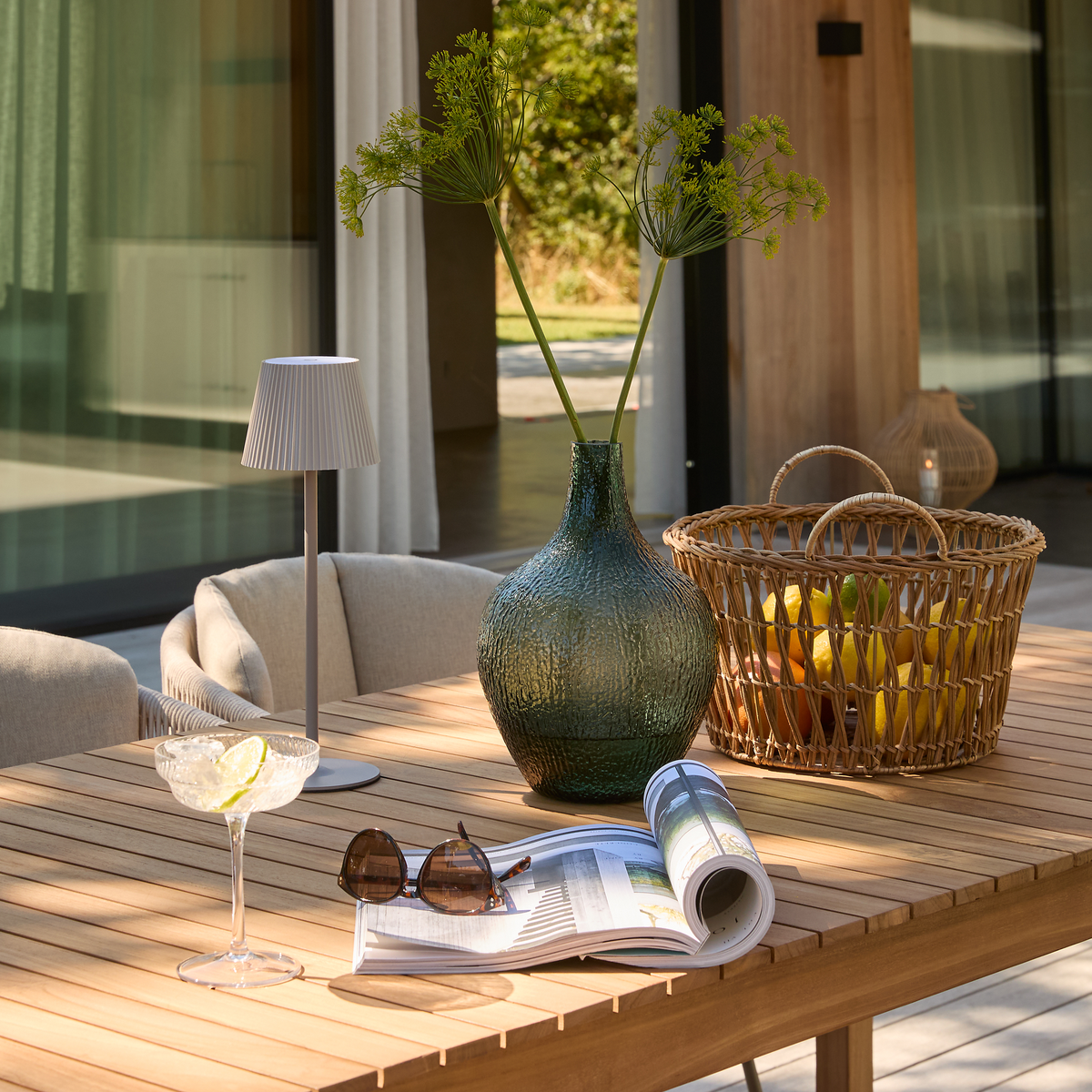 Træbord på terrasse med grøn glasvase med grene, kurv med frugt, bærbar bordlampe, cocktail med citronskive samt opslået magasin og solbriller i sollys.