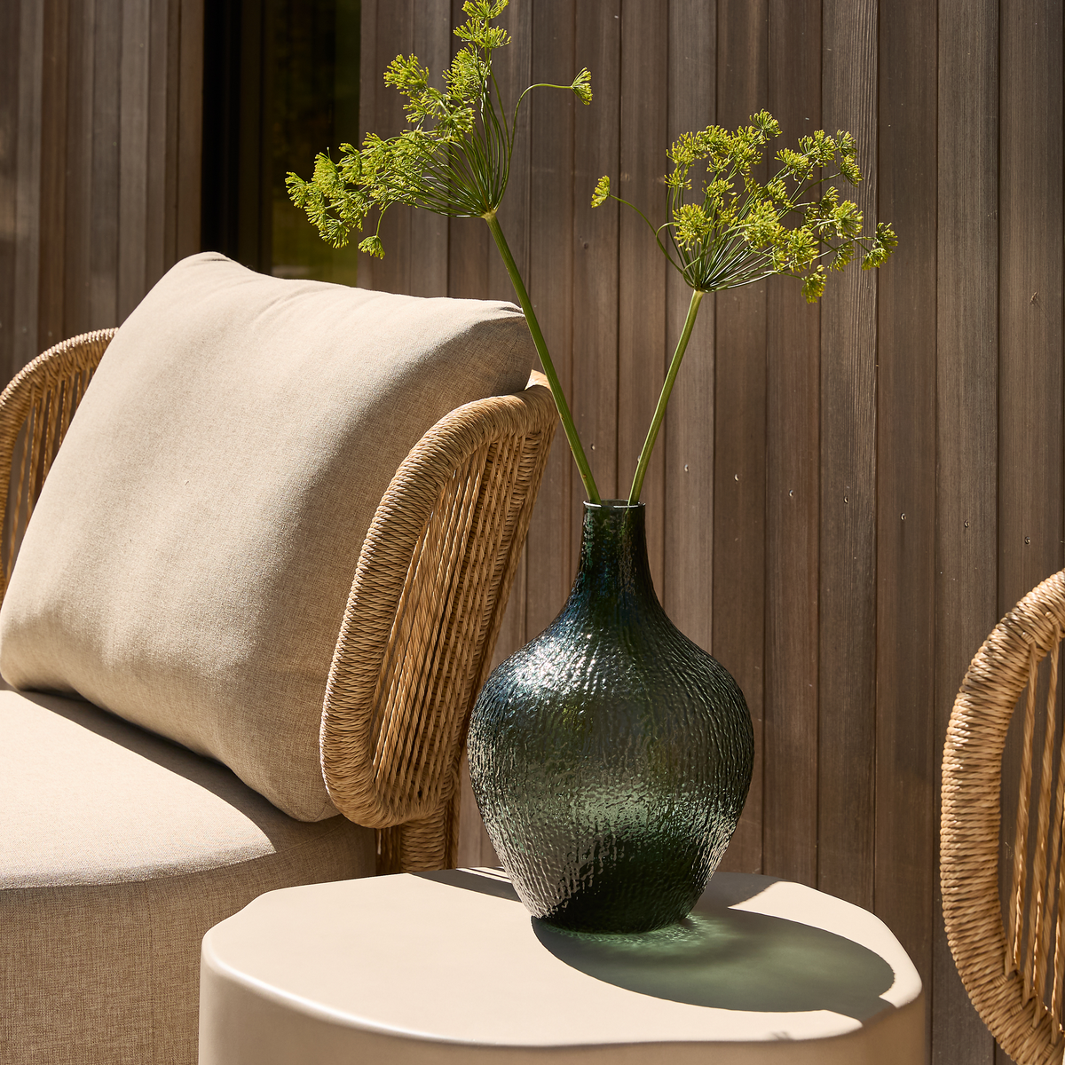 Grøn struktureret glasvase med dild‑lignende blomster på et lyst sidebord ved siden af en rattanstol, udendørs i sollys.