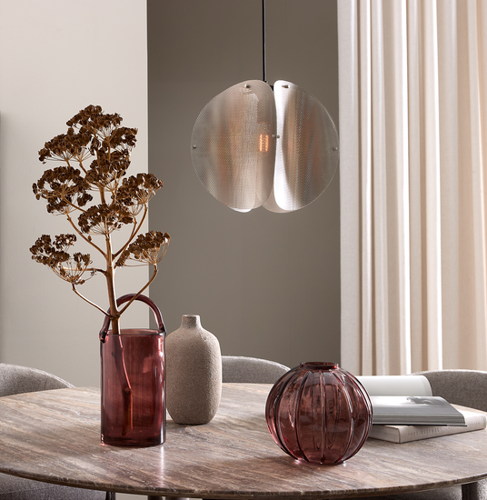 Rundt spisebord i træ med to vinrøde glasvaser, en beige keramikvase og en tørret gren; over bordet hænger en metallisk pendel, lyse gardiner i baggrunden.