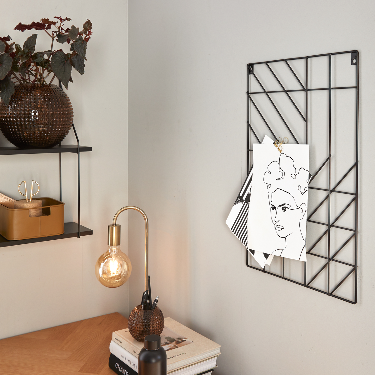 Hjemmekontorhjørne med træskrivebord, sort metalgitter med fastklemte illustrationer, tænd bordlampe i messing og glas, plante i brun vase på hylde og bøger på bordet med ryggene ”Chanel” og ”Kinfolk”.