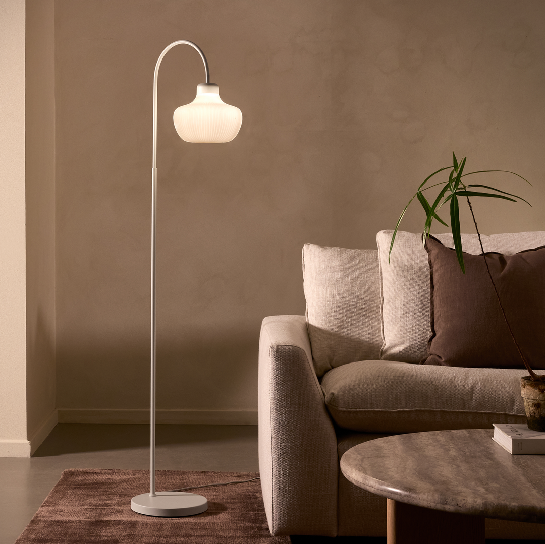 Hyggelig stue med tændt hvid gulvlampe ved siden af en beige sofa, brun pude, potteplante, bøger på et rundt stenslignende sofabord og brun tæppe i varm belysning.