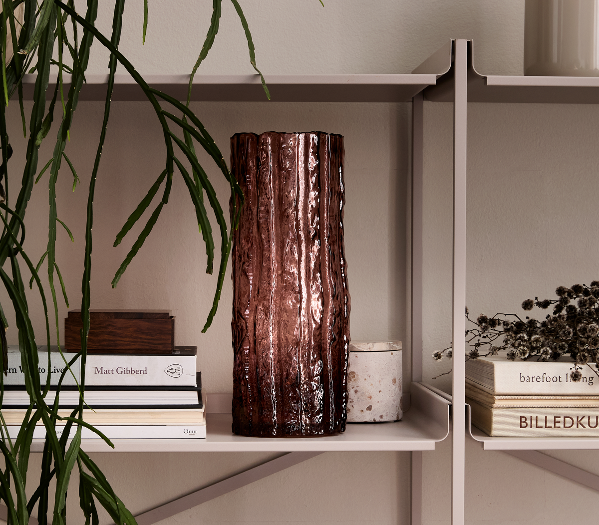 En beige reol med en rødbrun, riflede glasvase i midten, omgivet af en hængeplante og stakke bøger med titlerne Matt Gibberd, barefoot living, Ouur og BILLEDKU samt en lille lys beholder.