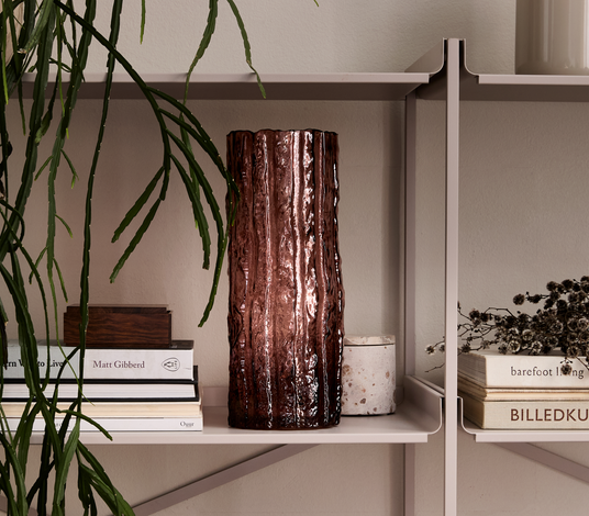 En beige reol med en rødbrun, riflede glasvase i midten, omgivet af en hængeplante og stakke bøger med titlerne Matt Gibberd, barefoot living, Ouur og BILLEDKU samt en lille lys beholder.