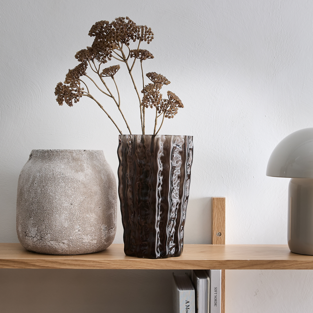 Brun bølget glasvase med tørrede blomster på en træhylde ved siden af en rustik krukke og en grå bordlampe.