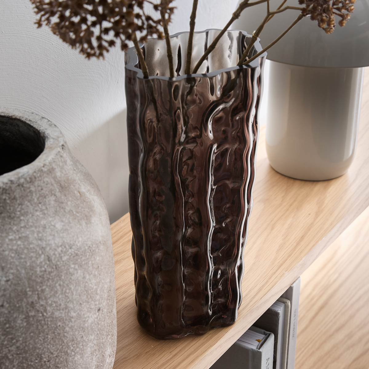 Brun, bølget glasvase på en træhylde med tørrede grene, ved siden af en grå lampe og en rå keramikvase.