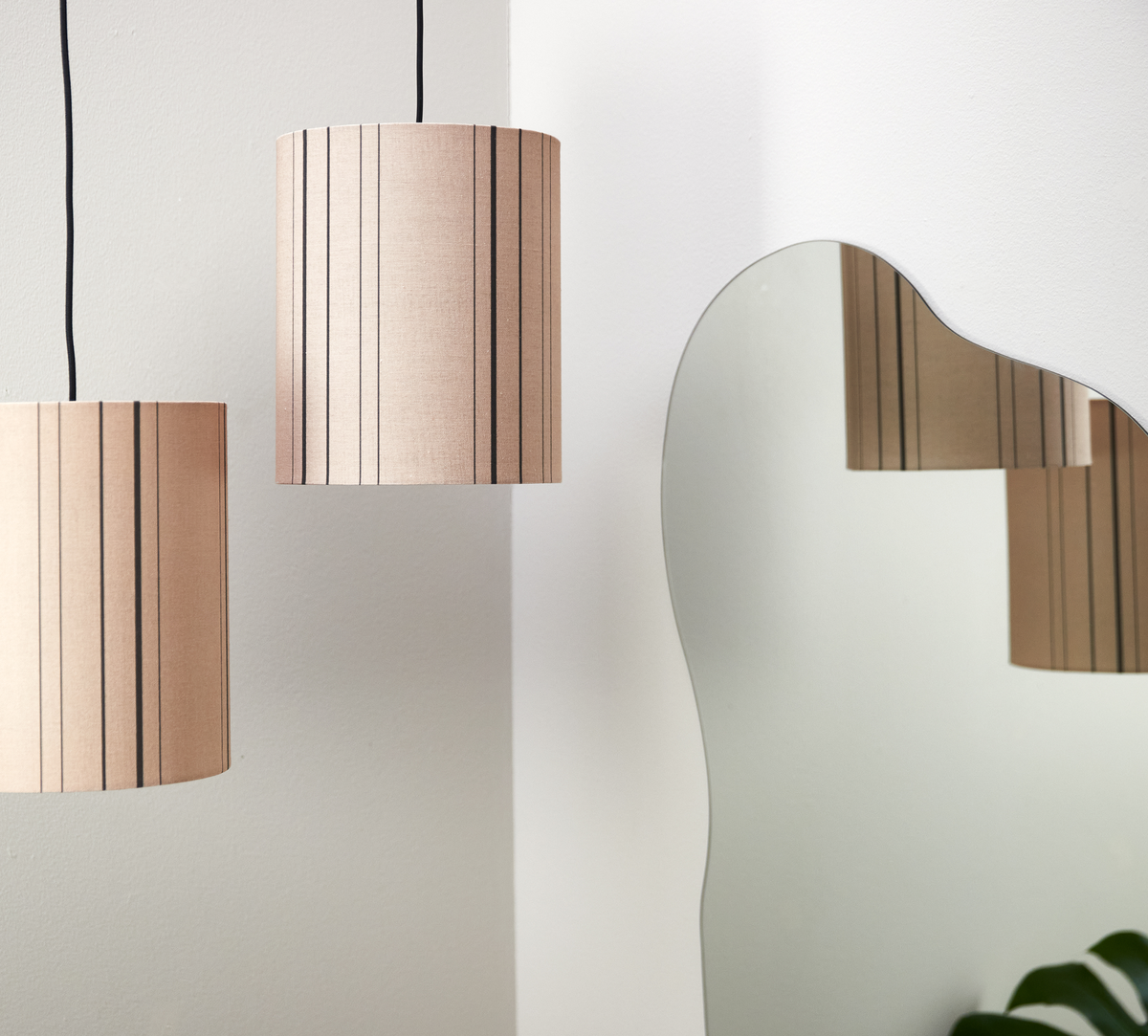 To beige, stribede pendler ved siden af et bølget spejl på en hvid væg, med lampens spejlbillede og et grønt blad i hjørnet.