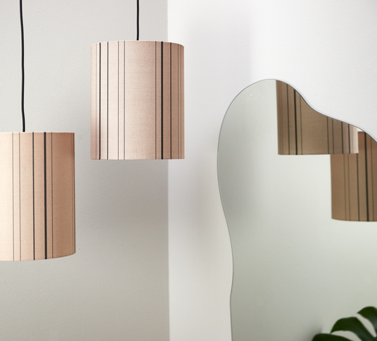 To beige, stribede pendler ved siden af et bølget spejl på en hvid væg, med lampens spejlbillede og et grønt blad i hjørnet.