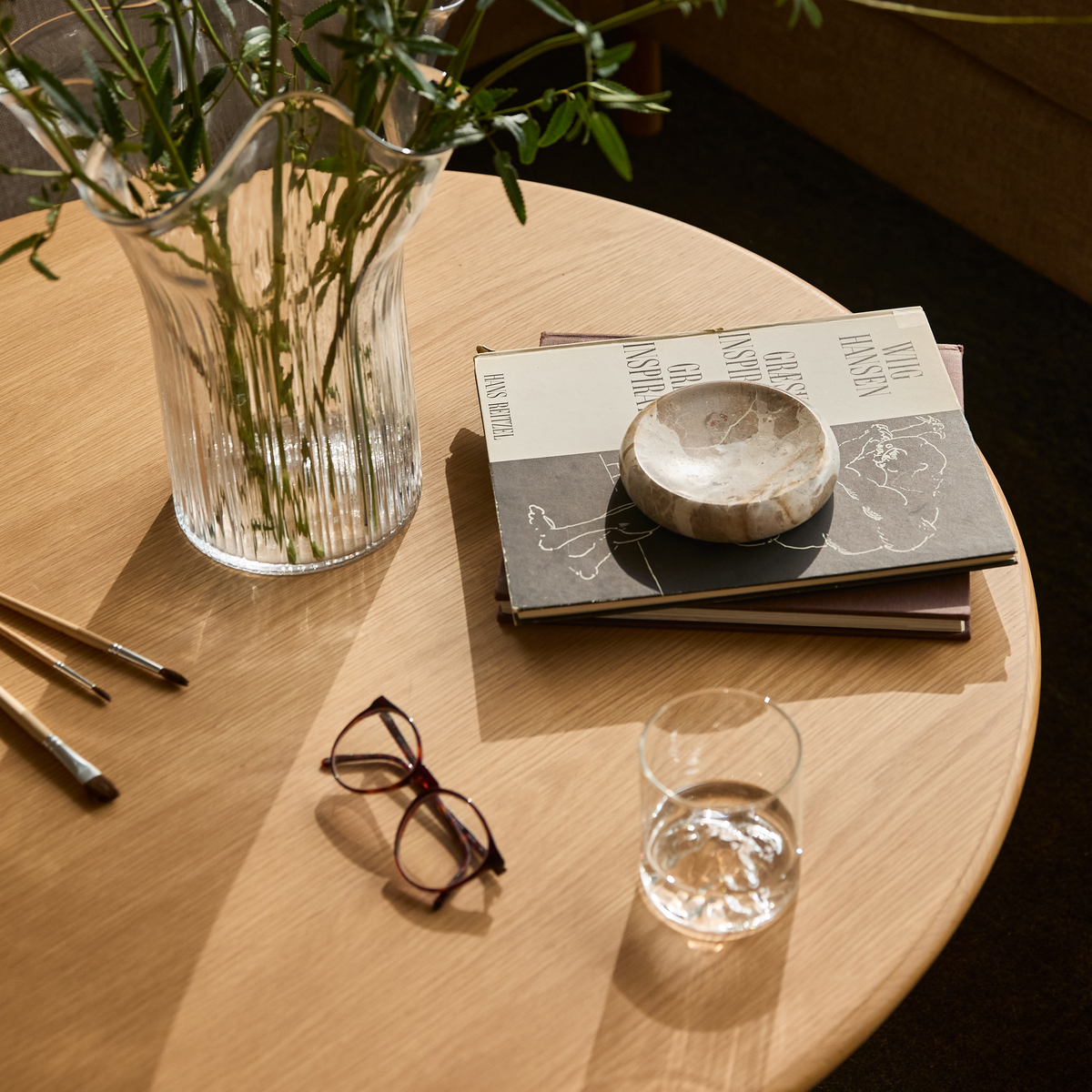Solbelyst rundt sofabord i lyst træ med rillet glasvase med grønne kviste, stak designbøger (VING HANSEN), lille skål i sten, vandglas, pensler og et par briller.
