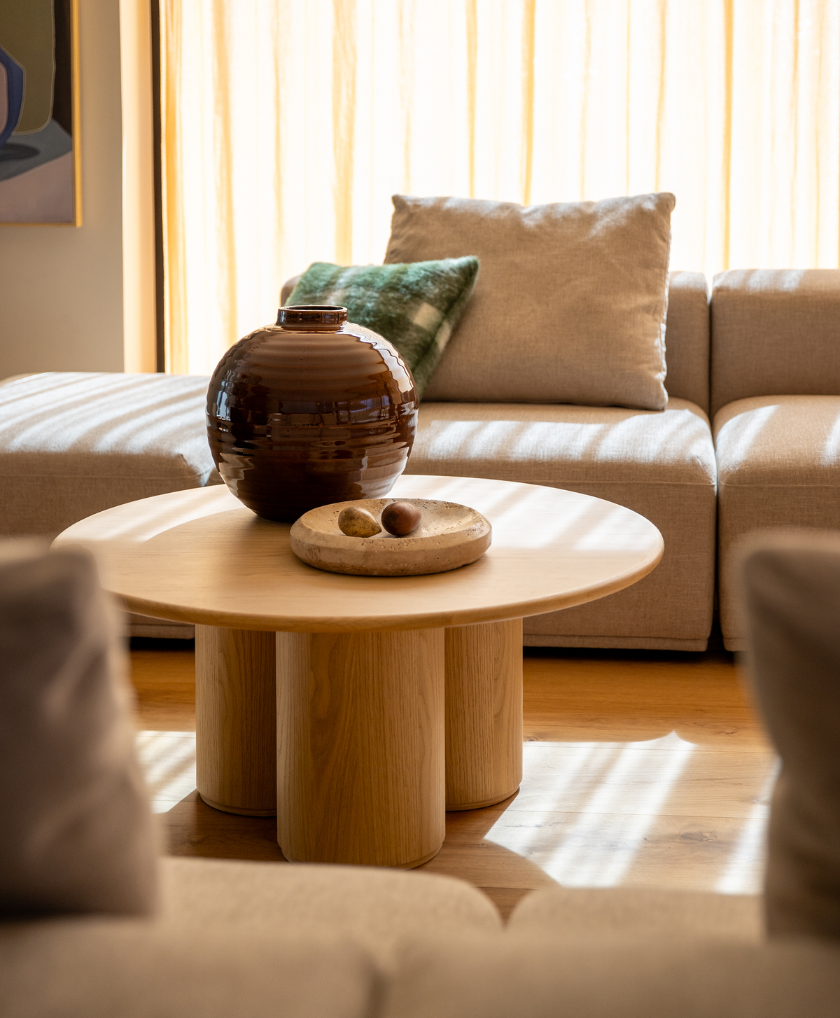 Beige sofa og rundt sofabord i lyst træ med brun keramikvase og skål, solstriber gennem lyse gardiner.