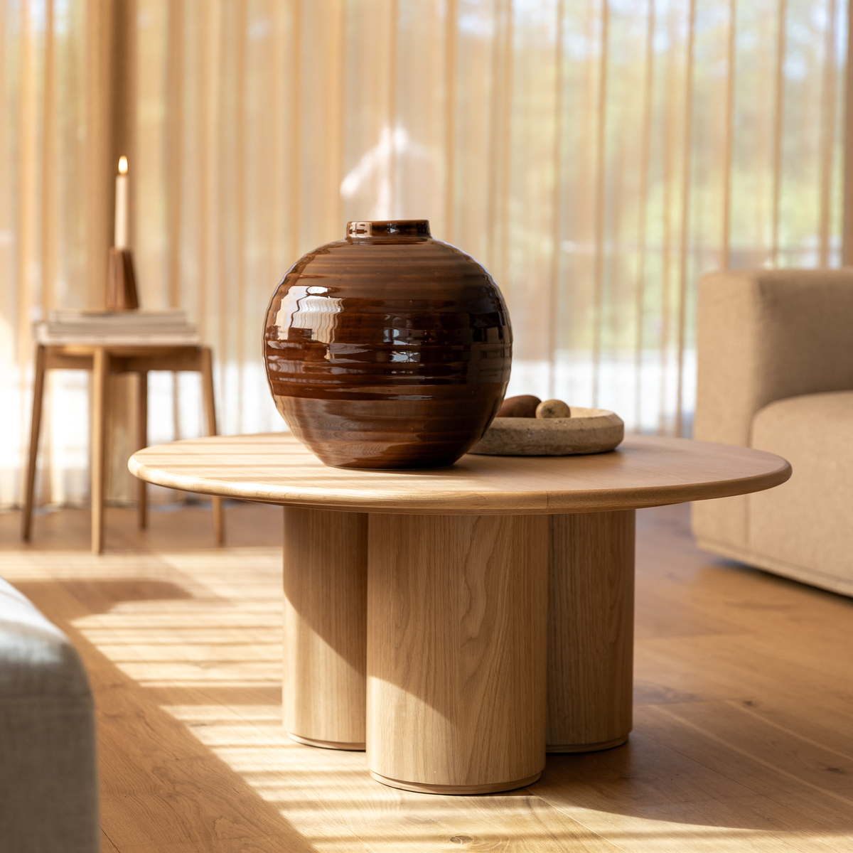 Rundt sofabord i eg med cylindriske ben, brun keramisk vase ovenpå; blødt dagslys gennem transparente gardiner, beige sofa i baggrunden.