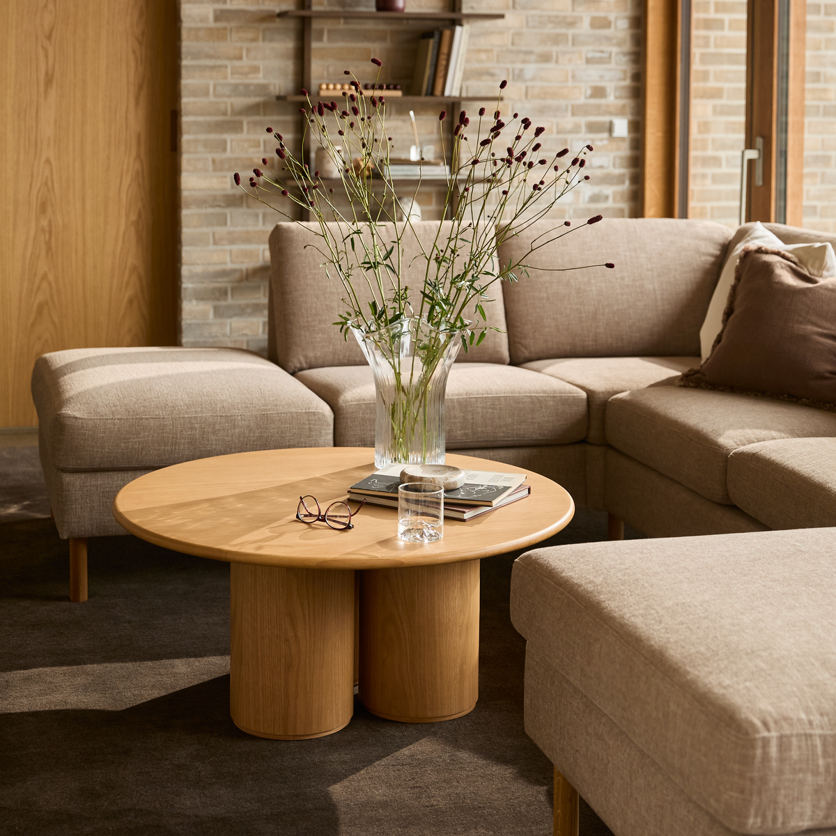 Beige hjørnesofa med rundt sofabord i træ, vase med blomster, bøger og et glas i en lys stue.