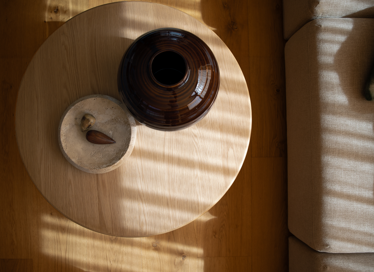 Top-down visning af et rundt sofabord i lyst egetræ med en mørkebrun keramisk vase og et lille stentallerken med to små objekter; sollys gennem persienner laver striber ved siden af en beige sofa.