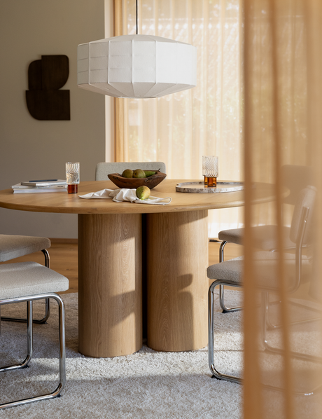 Rundt eget spisebord med cylindriske ben, polstrede stole med kromben og en hvid pendellampe over; lette gardiner, frugtskål med pærer, glas og bakke på bordet i et lyst spisestue-miljø.