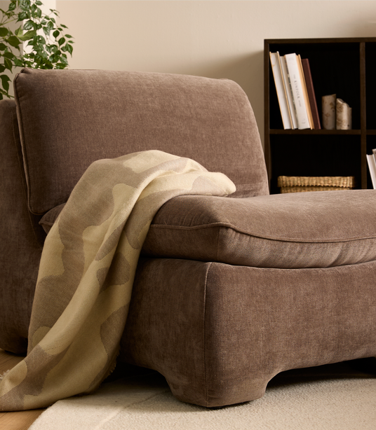 Brun loungestol i blødt tekstil med beige plaid over armlænet, ved siden af reol og plante i en rolig stue.