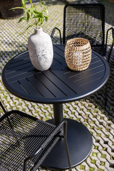 Rundt sort udendørsbord med slidsede lameller, en grå vase med grøn kvist og en flettet lanterne med hvidt lys på en brolagt terrasse i sollys med sorte stole.
