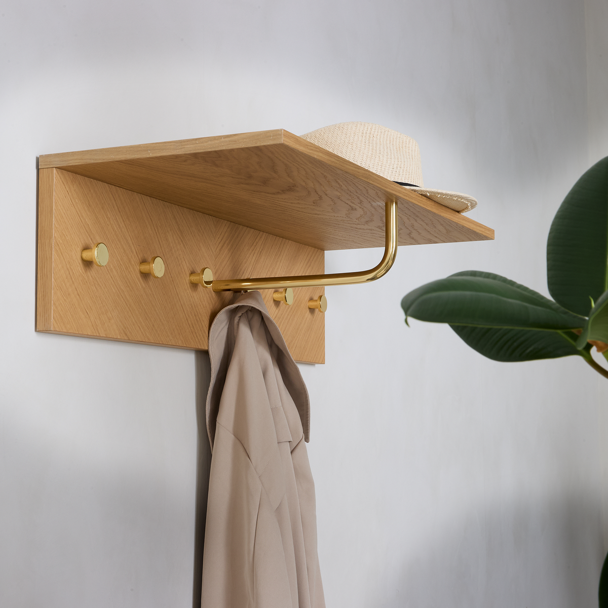 Væghængt hattehylde i eg med messingfarvede knager og bøjlestang; en beige frakke hænger, og en stråhat ligger på hylden, med en grøn plante ved siden af.