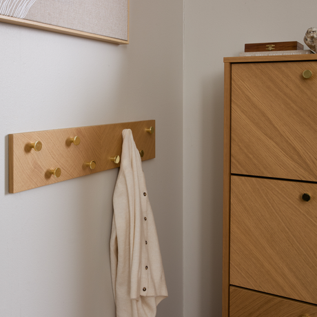 Vægmonteret egetræsskinne med messingknopper, en beige cardigan hænger på en krog; lys trækommode ved siden af i en minimalistisk entré.