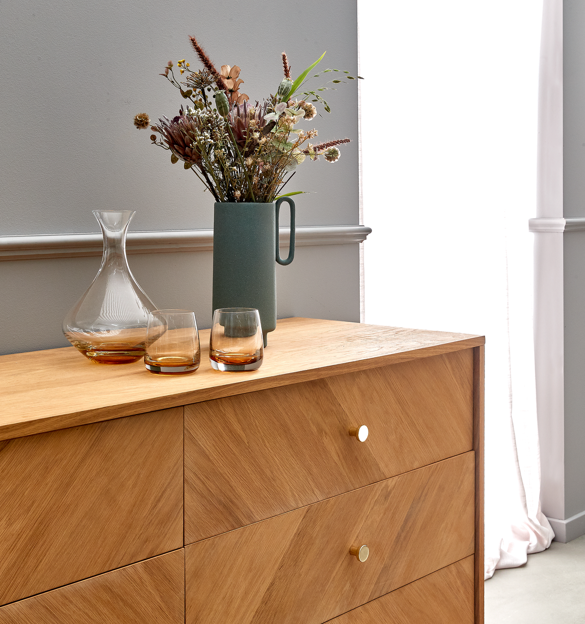 Træskænk med sildebensmønster og messingknopper, pyntet med grøn vase med tørrede blomster, en glaskaraffel og to ravfarvede glas ved et lyst vindue.