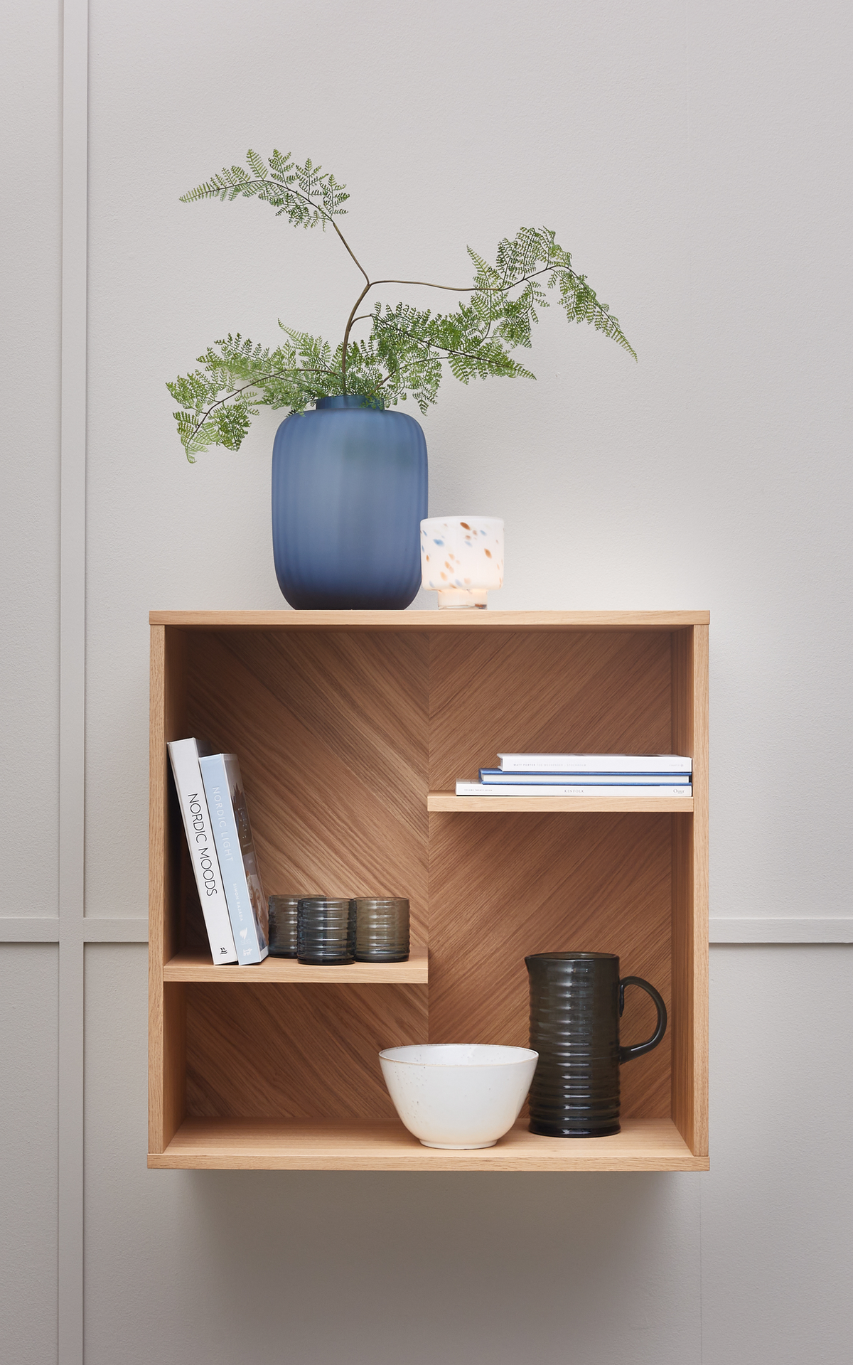 Vægmonteret hylde i lyst træ med blå vase og grønne grene, lille hvid lysestage, skål, kande og bøger, hvoraf en har titlen NORDIC MOODS.