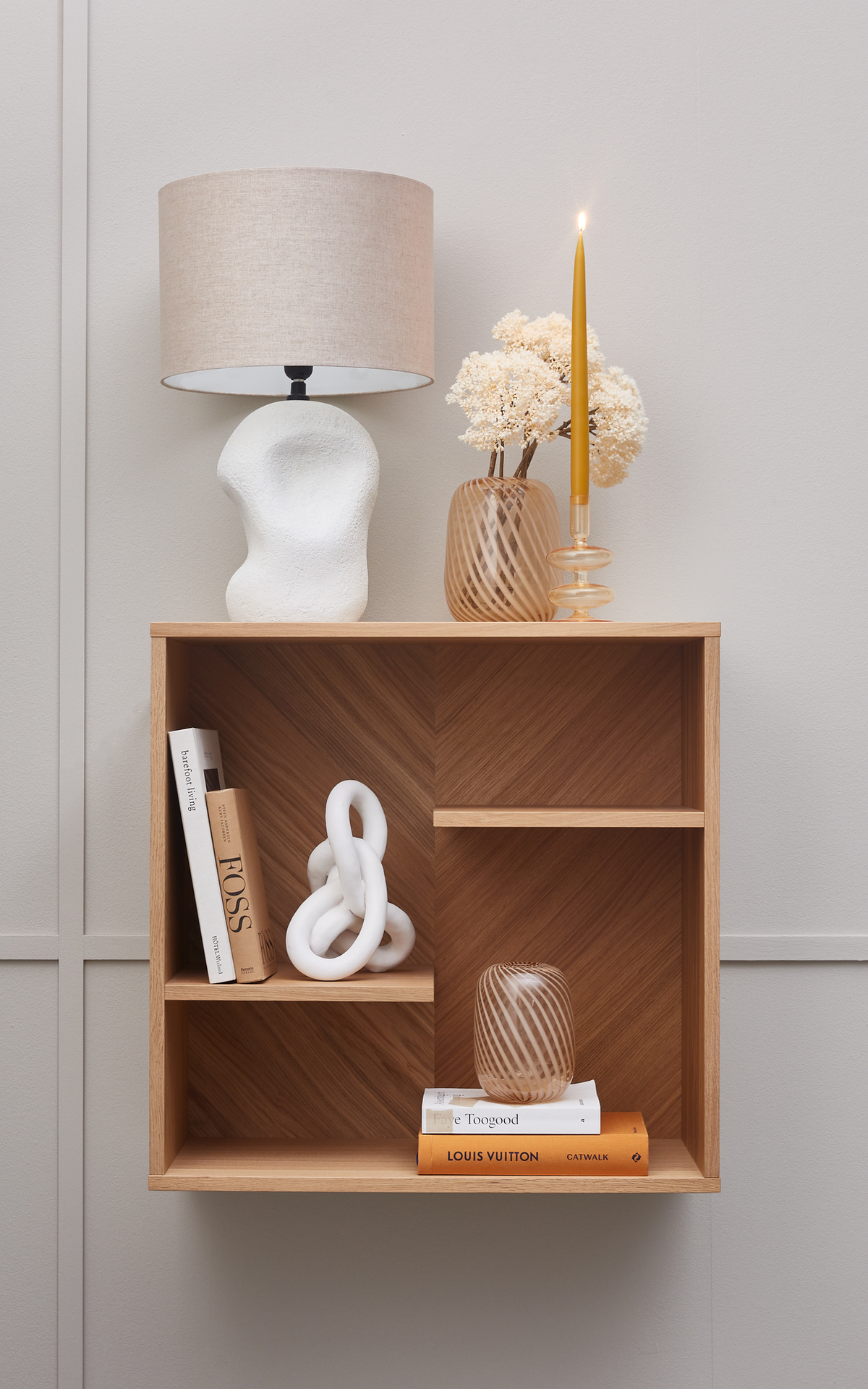 Minimalistisk væghylde i træ med bøger (“Faye Toogood”, “Louis Vuitton Catwalk”, “FOSS”), skulpturel hvid dekoration, beige bordlampe, stribet glasvase med tørrede blomster og et tændt ravfarvet stagelys.