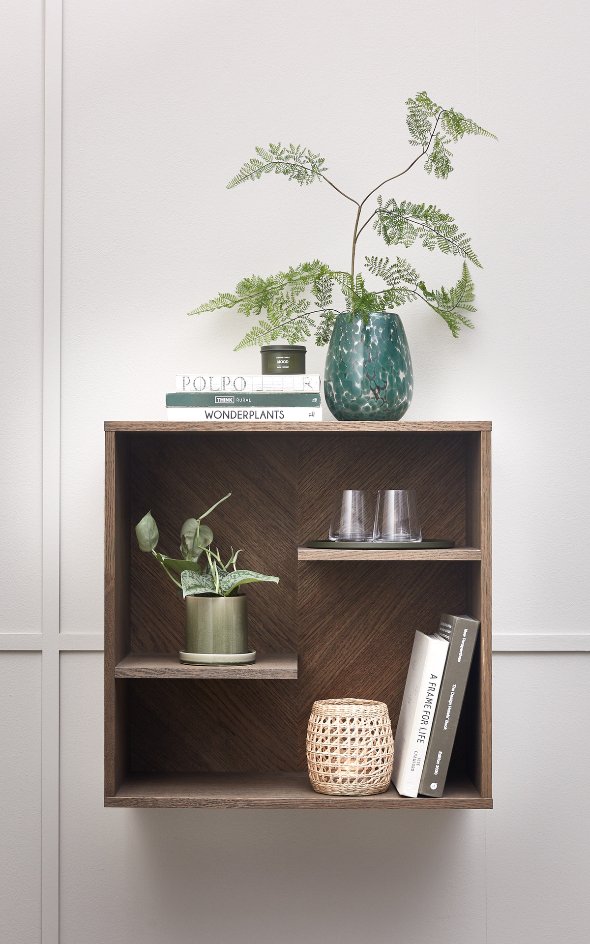 Vægmonteret reol i mørkt træ med bøger (WONDERPLANTS, POLPO, A FRAME FOR LIFE), grønne planter i potte og grøn glasvase, to glas og en flettet lille lanterne mod en lys væg.