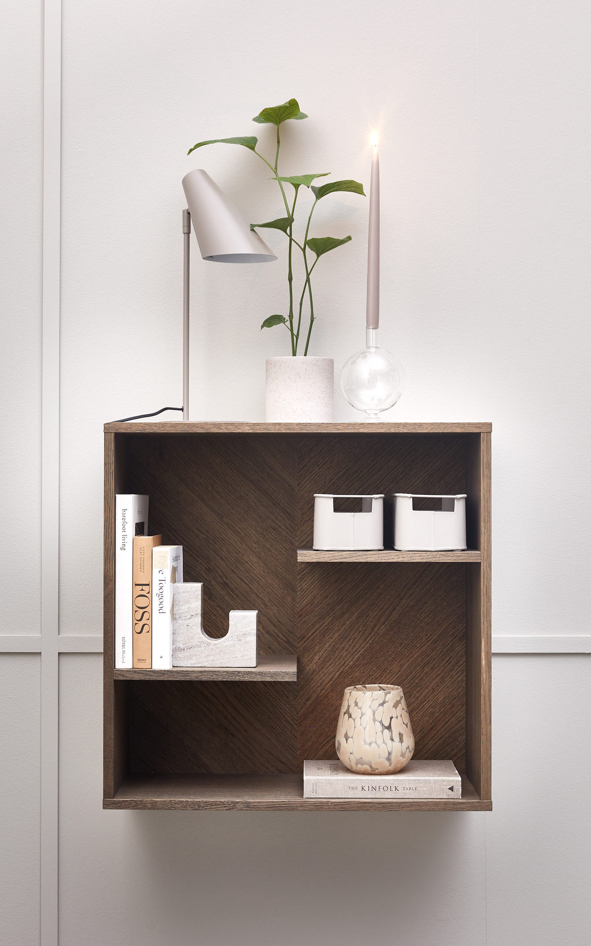 Væghængt træhylde med bøger ("FOSS", "The Kinfolk Table"), marmorbogstøtte, mønstret vase, to hvide opbevaringsbokse, lille potteplante, moderne bordlampe og et tændt stagelys mod en lys væg.