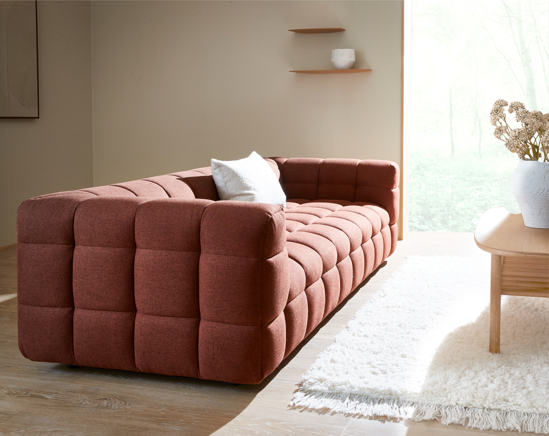 Rustfarvet, dybt polstret sofa i stue med hvidt shag-tæppe og træbord i dagslys.