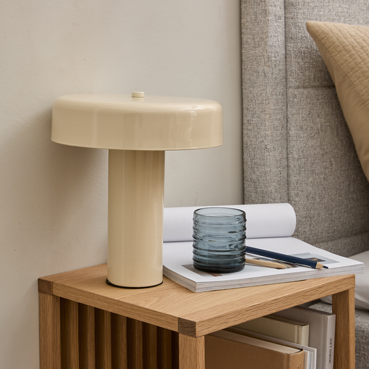 Beige bordlampe på et træ-natbord ved siden af en grå sengegavl, med et blåt glas og en notesbog med blyanter i blødt dagslys.