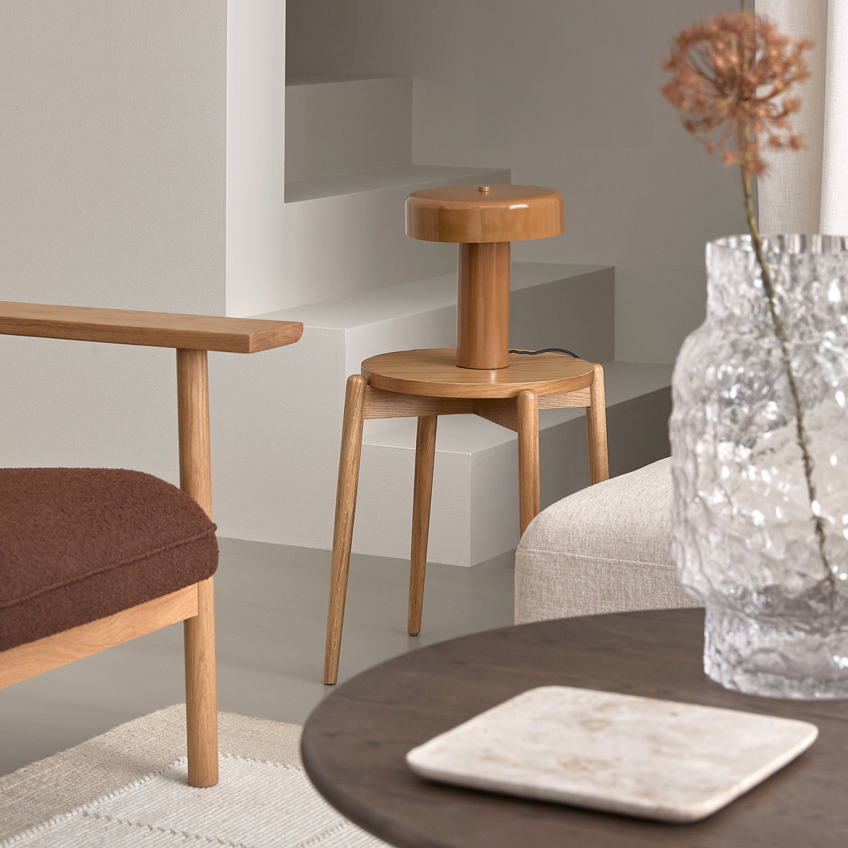 Lyst, minimalistisk hjørne af stuen med trælænestol, lille rundt sidebord med karamelfarvet bordlampe, tekstureret glasvase og mørkt sofabord i neutrale nuancer.