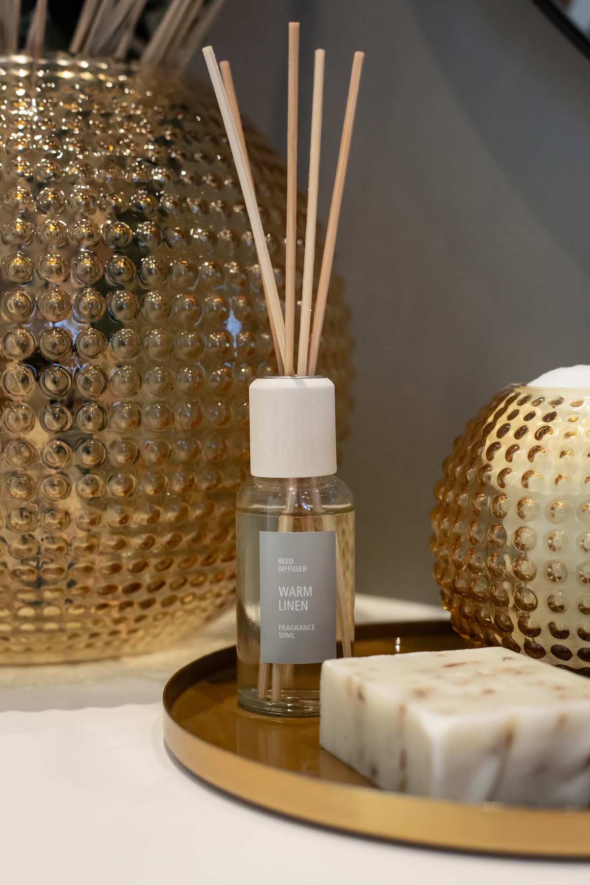 Nærbillede af en duftspreder med træpinde på en gylden bakke ved siden af teksturerede glasvaser og sæbe; etiketten viser REED DIFFUSER WARM LINEN FRAGRANCE 50ML.