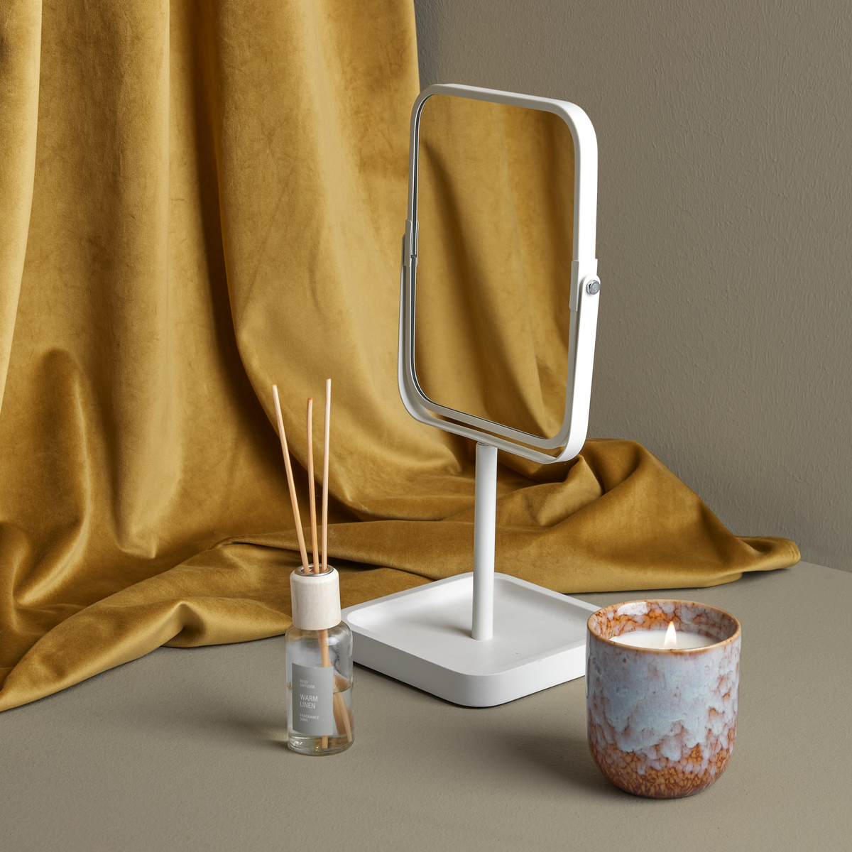 Rektangulært hvidt bordspejl med bakke ved siden af et tændt keramisk duftlys og duftpinde mærket ”Urban Linen”, foran en sennepsgul fløjlbaggrund.