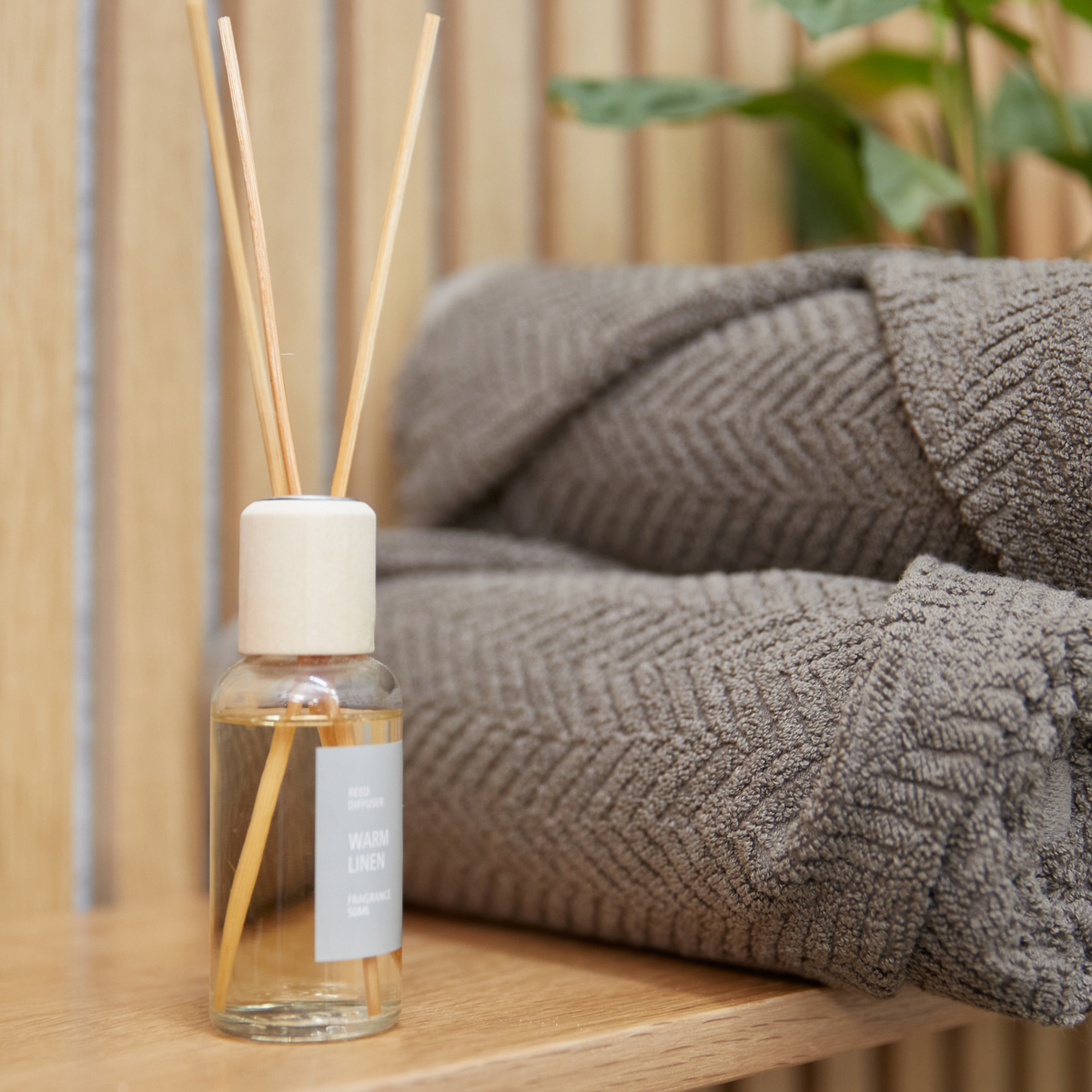 Nærbillede af duftpinde i glasflaske med etiketten REED DIFFUSER WARM LINEN FRAGRANCE ved siden af foldede grå håndklæder på en træhylde.