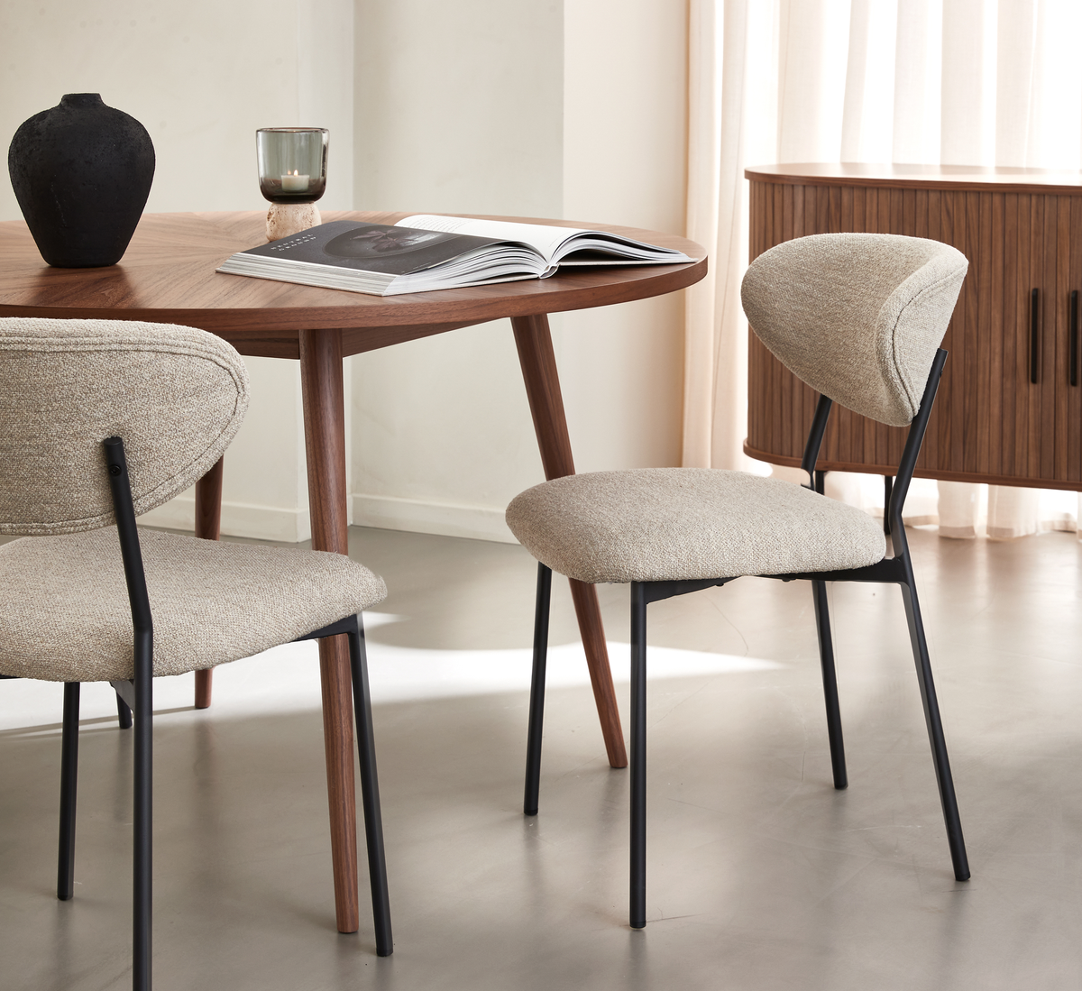 Beige polstrede spiseborde med sorte metalben omkring et rundt valnødsbord med opslået bog, lys i glas og sort vase i et lyst, minimalistisk spisestue.