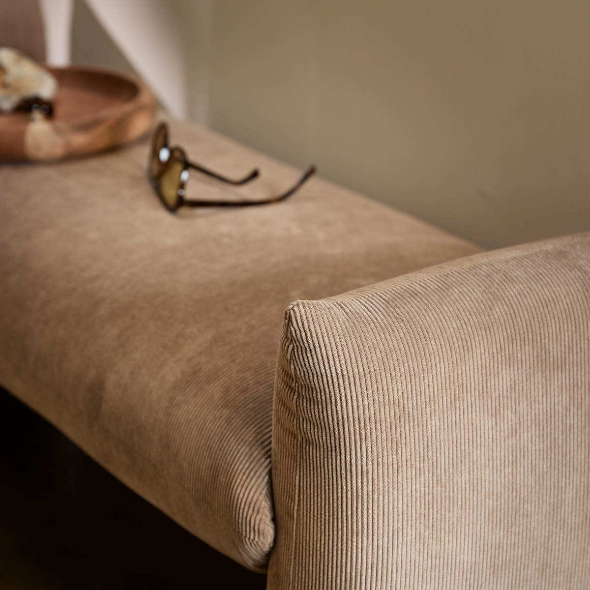 Nærbillede af beige, riflet polstret bænk/sofa med solbriller og en træskål på sædet.