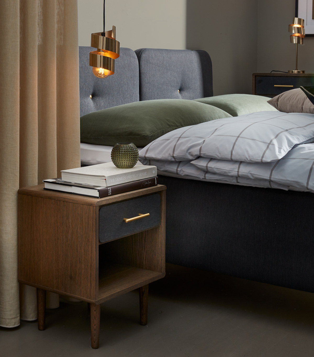 Soveværelse med mørkeblåt polstret sengegærde, træ-natbord med messinggreb, to bøger inkl. THE DESIGN HOTELS BOOK, lille grøn glasvase og hængende messingpendel; grønne puder og ternet dynebetræk.