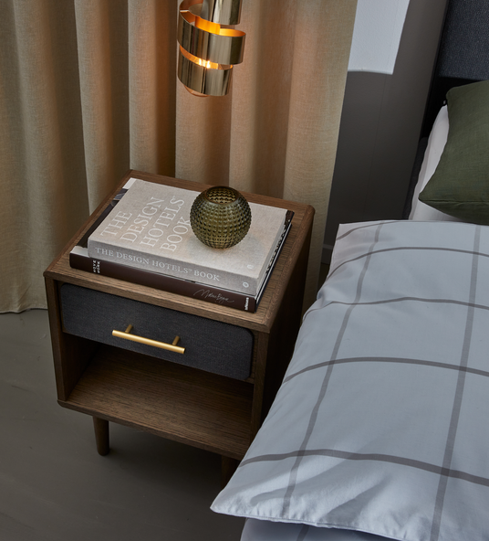 Natbord i mørkt træ med messinggreb ved siden af sengen; ovenpå ligger designbøger med titlen THE DESIGN HOTELS BOOK og en lille rillet glasvase, med messingpendel og beige gardiner i baggrunden.