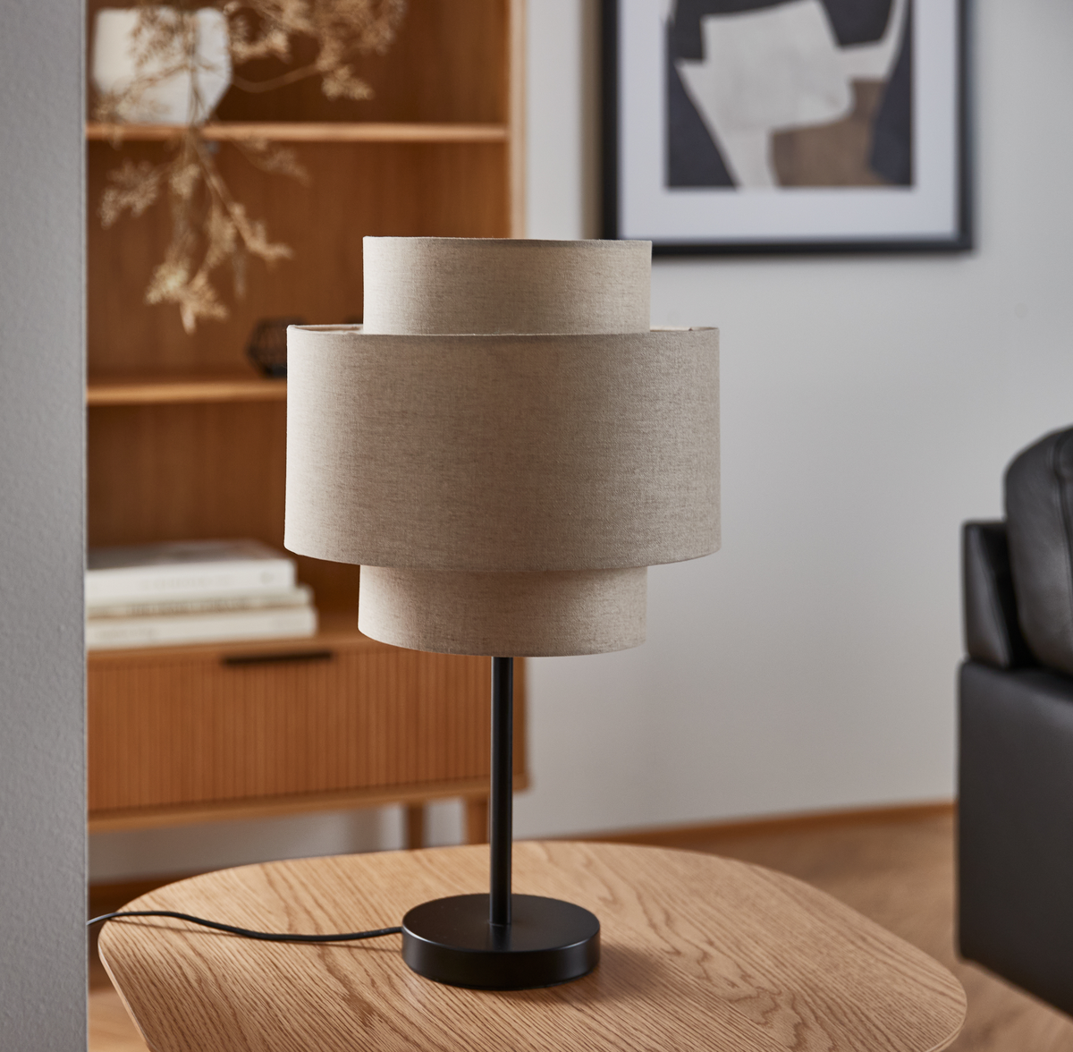 Bordlampe med lagdelt beige tekstilskærm og sort fod på et træbord i en stue med reol og abstrakt billede.