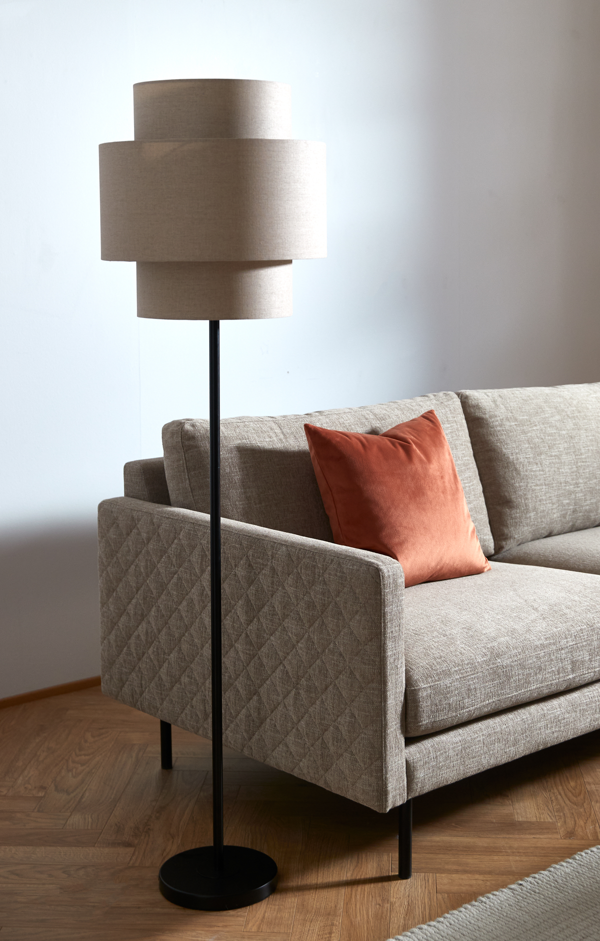 Beige gulvlampe med lagdelt lampeskærm ved siden af beige sofa med rustfarvet pude på trægulv i en lys stue.