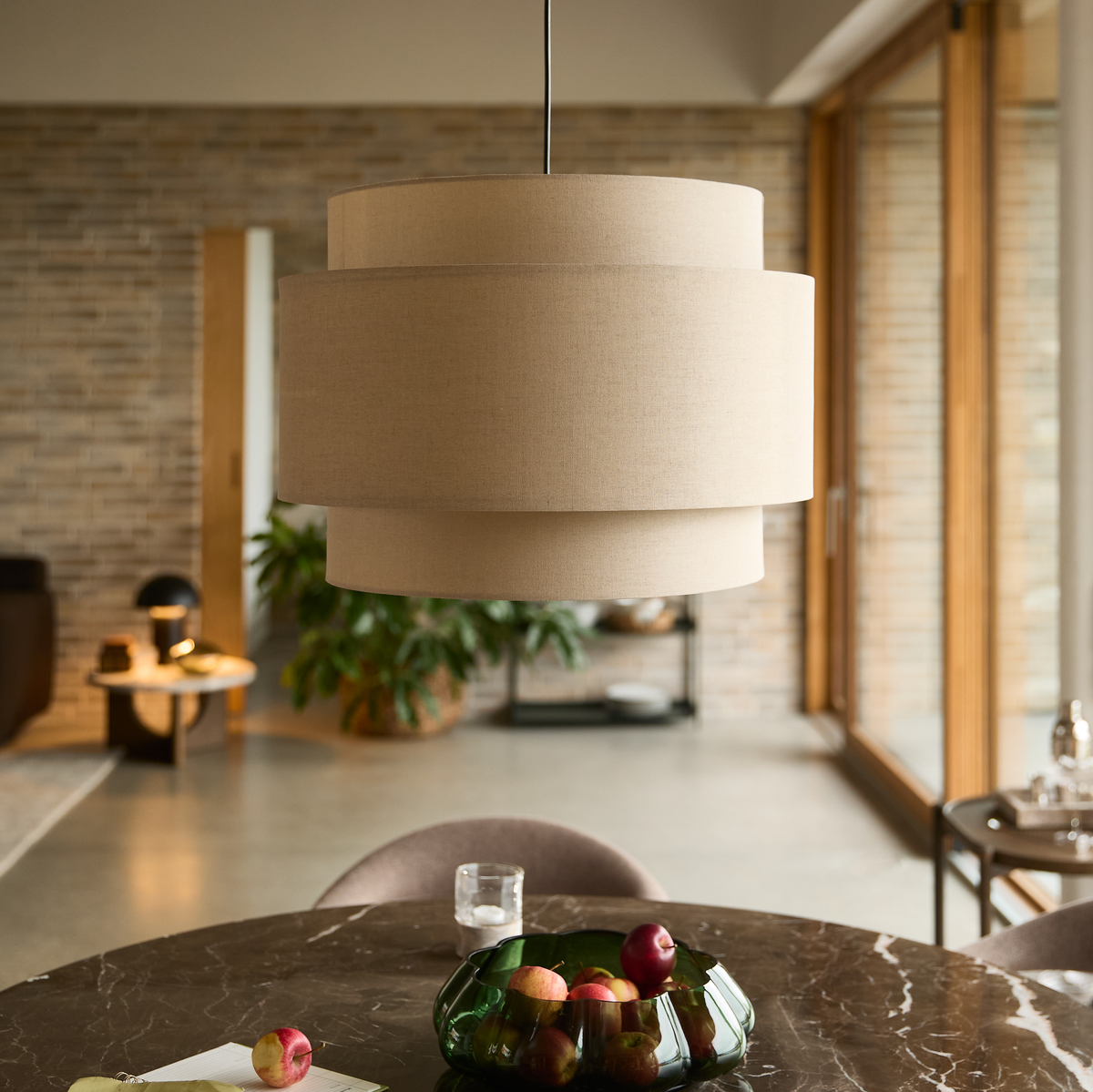 Beige pendellampe over et marmorbord med grøn glasskål med frugt i en lys, moderne spisestue.