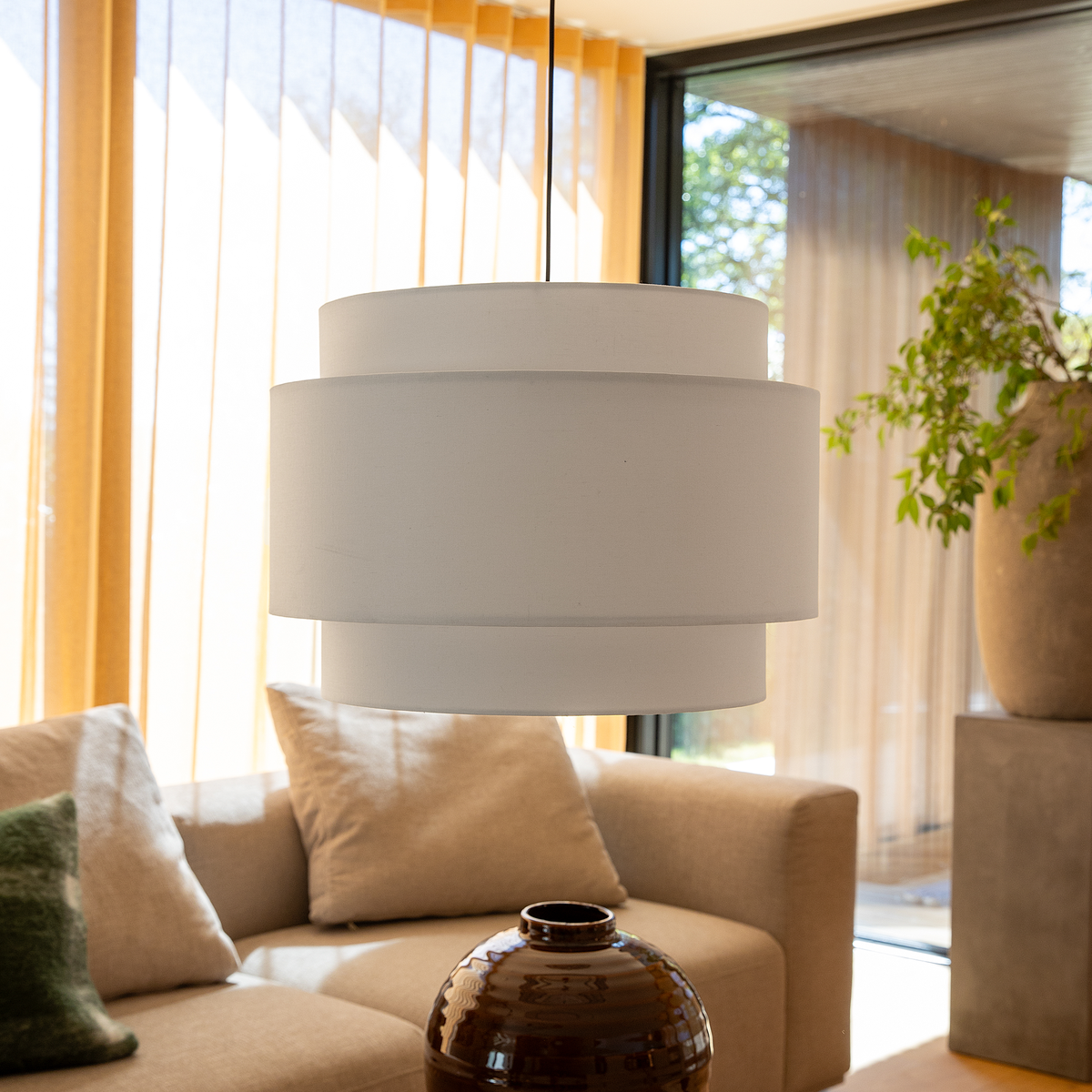 Beige stue med stor hvid lagdelt pendellampe over sofa, keramikvase på bord, lyse trælameller og grønne planter i dagslys.