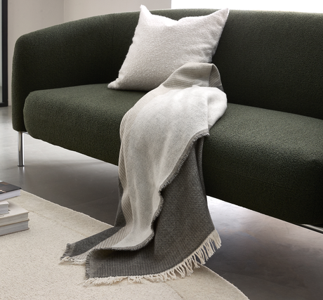 Mørkegrøn sofa med lys pude og tofarget grå plaid med frynser draperet over sædet i en minimalistisk stue.