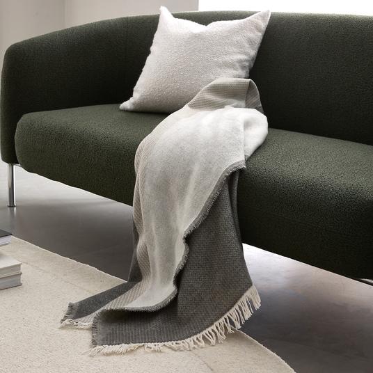 Mørkegrøn sofa med hvid pude og gråhvid plaid med frynser lagt over kanten, på et lyst tæppe i en lys stue.