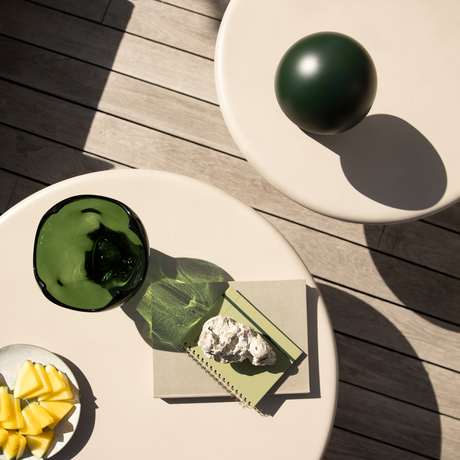 Overblik over to beige runde borde på en træterrasse med en mørkegrøn dekorationskugle, en grøn glasbeholder der kaster skygge, to notesbøger med en sten ovenpå og en lille tallerken med ananasstykker i sollys.