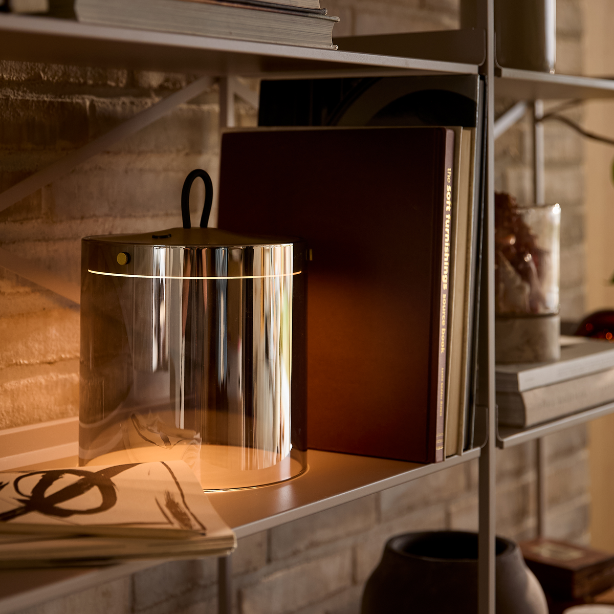 Nærbillede af en reol med en spejlblank bordlampe, der giver varmt lys ved siden af en brun bog med titlen Soft Furnishings Source Book, samt bøger og dekor i en hyggelig stue.