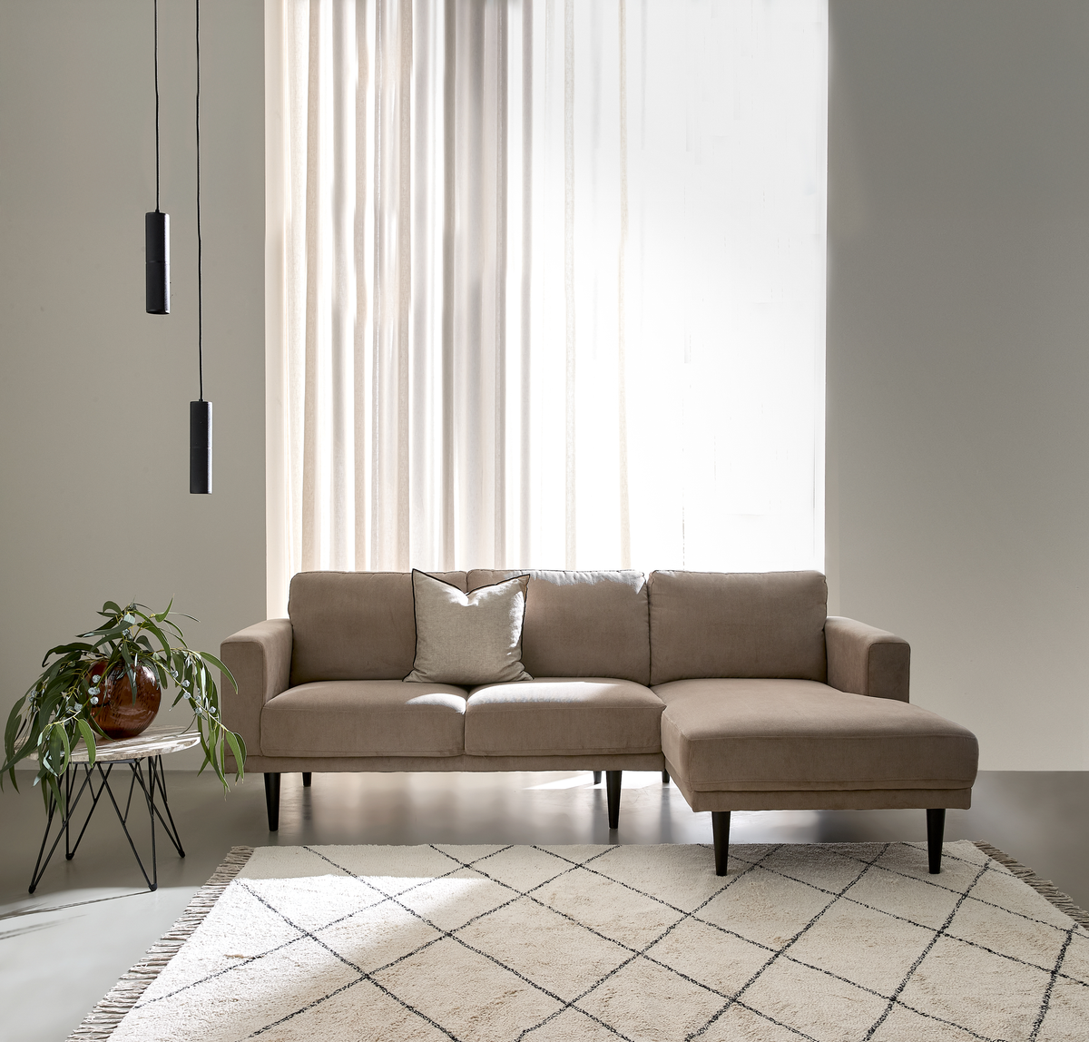 Beige hjørnesofa med chaiselong i en lys, minimalistisk stue med lette gardiner, to sorte pendler, et lille sidebord med grøn plante og et lyst tæppe med rudemønster.