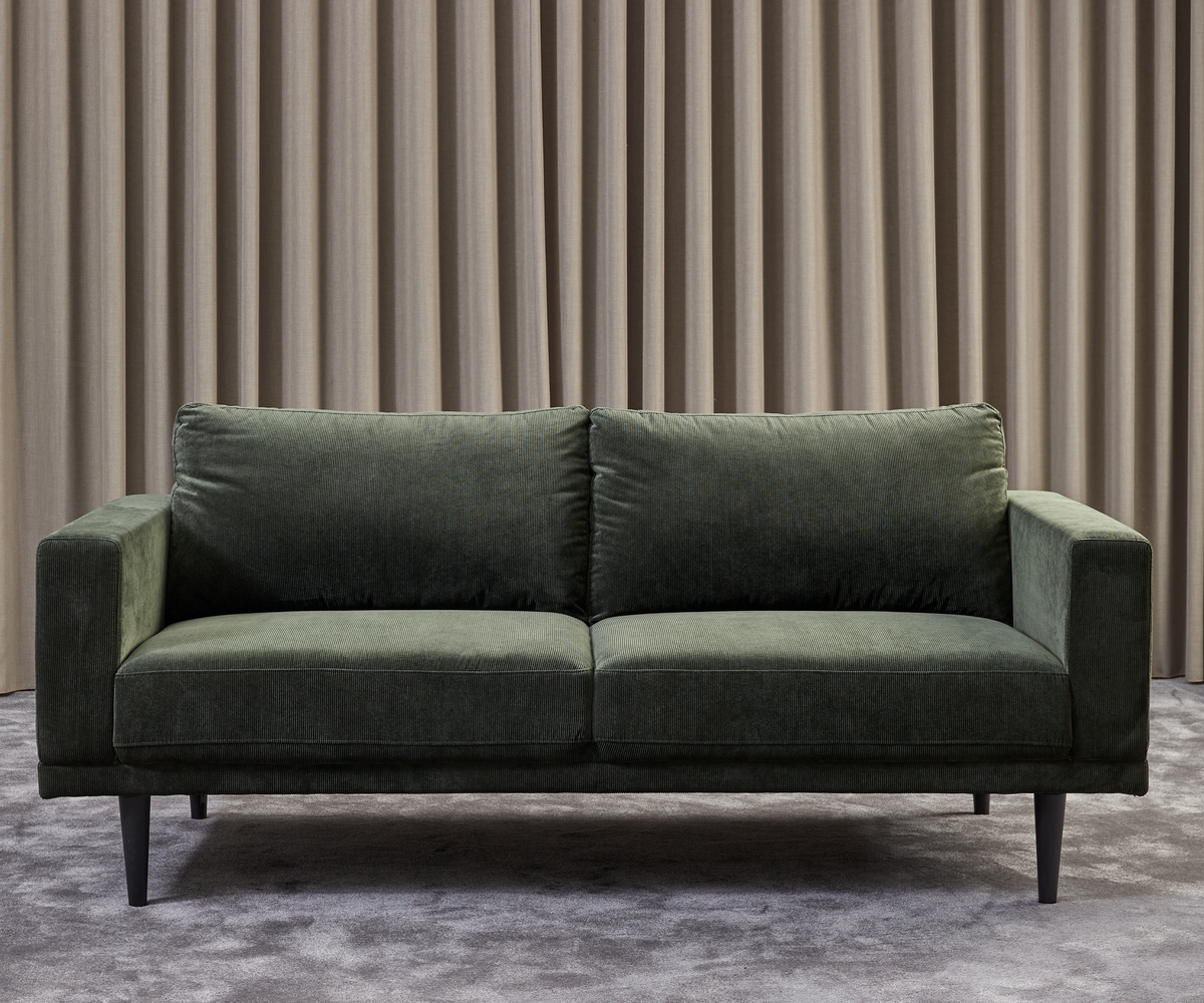 Mørkegrøn tosæders sofa med slanke sorte ben foran beige plisserede gardiner på gråt tæppe.