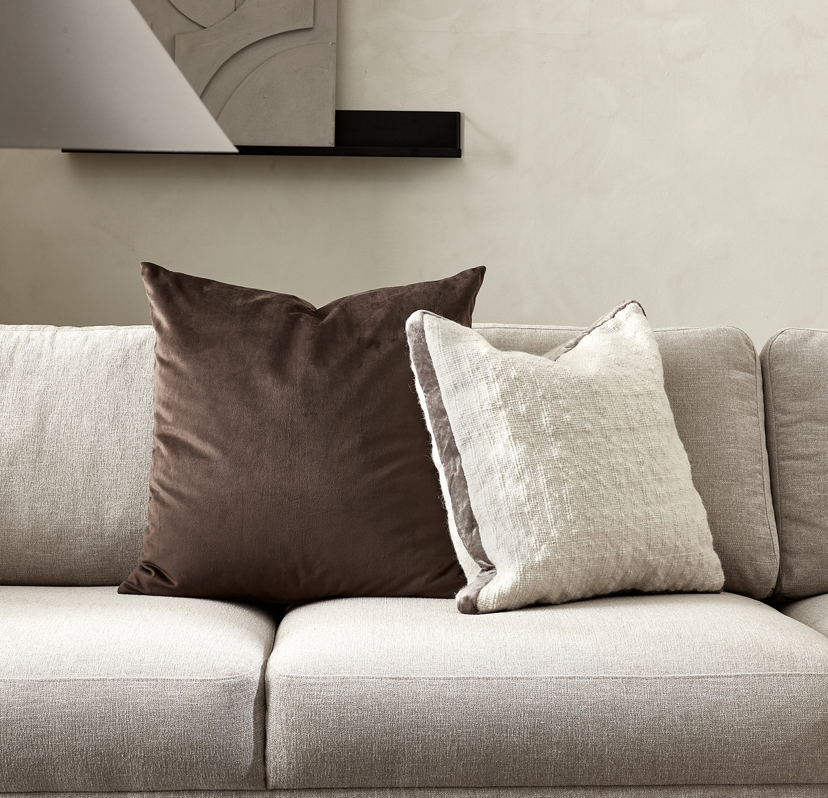 To pyntepuder, en brun og en lys beige, på en lysegrå sofa i et minimalistisk stue-miljø.