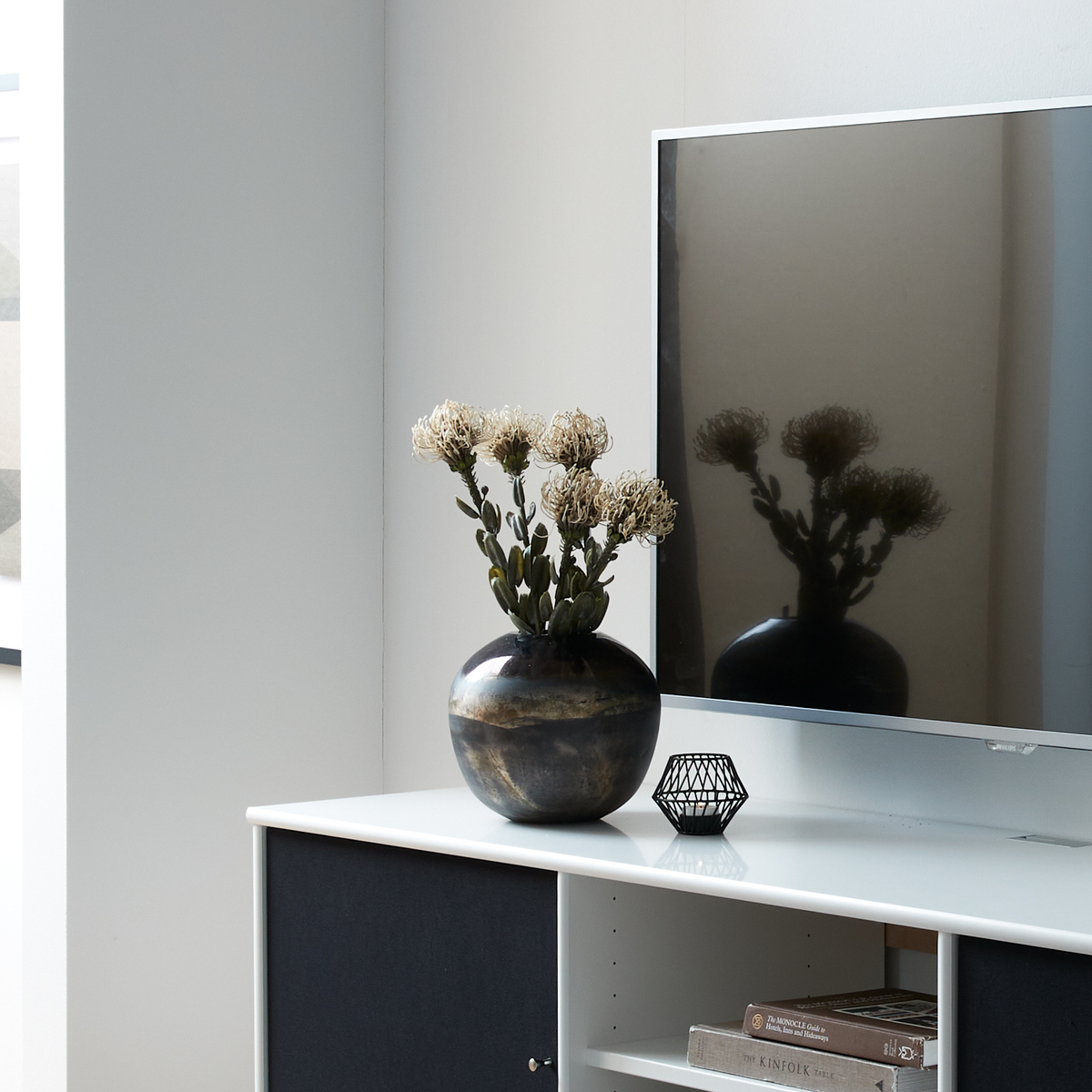 TV på hvid skænk med rund keramisk vase med tørrede blomster, geometrisk fyrfadsstage og bøger, hvoraf én har titlen Kinfolk.